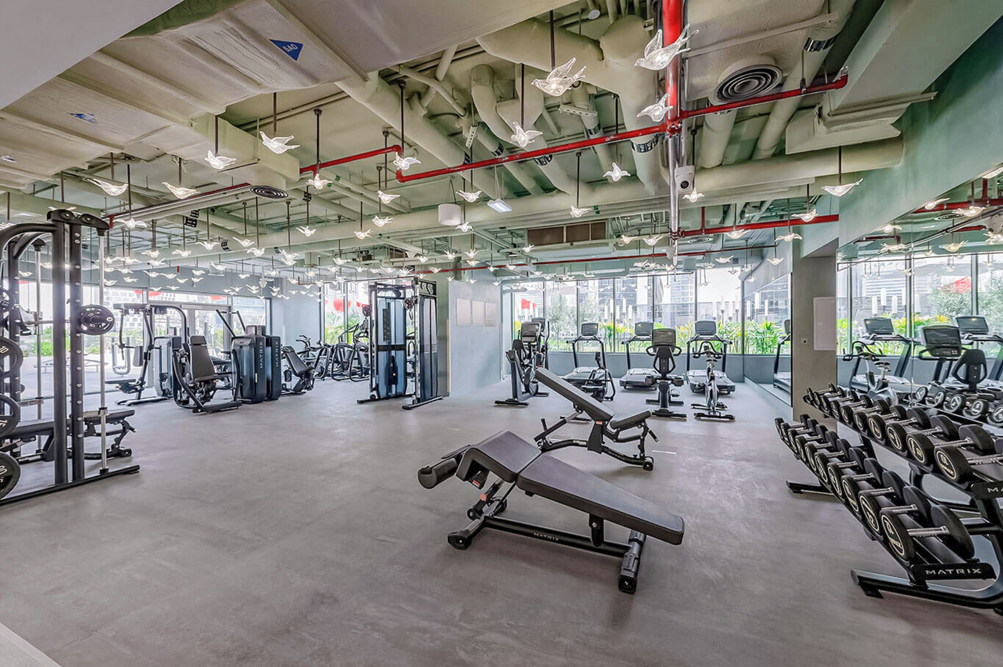 A spacious gym with various exercise equipment, large windows, and a modern industrial ceiling design.