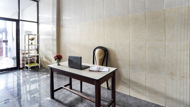 Un hall d’hôtel moderne avec une entrée dotée d’une porte en verre, un bureau en bois avec un ordinateur portable et un vase de fleurs, et une chaise placée contre un mur à texture.