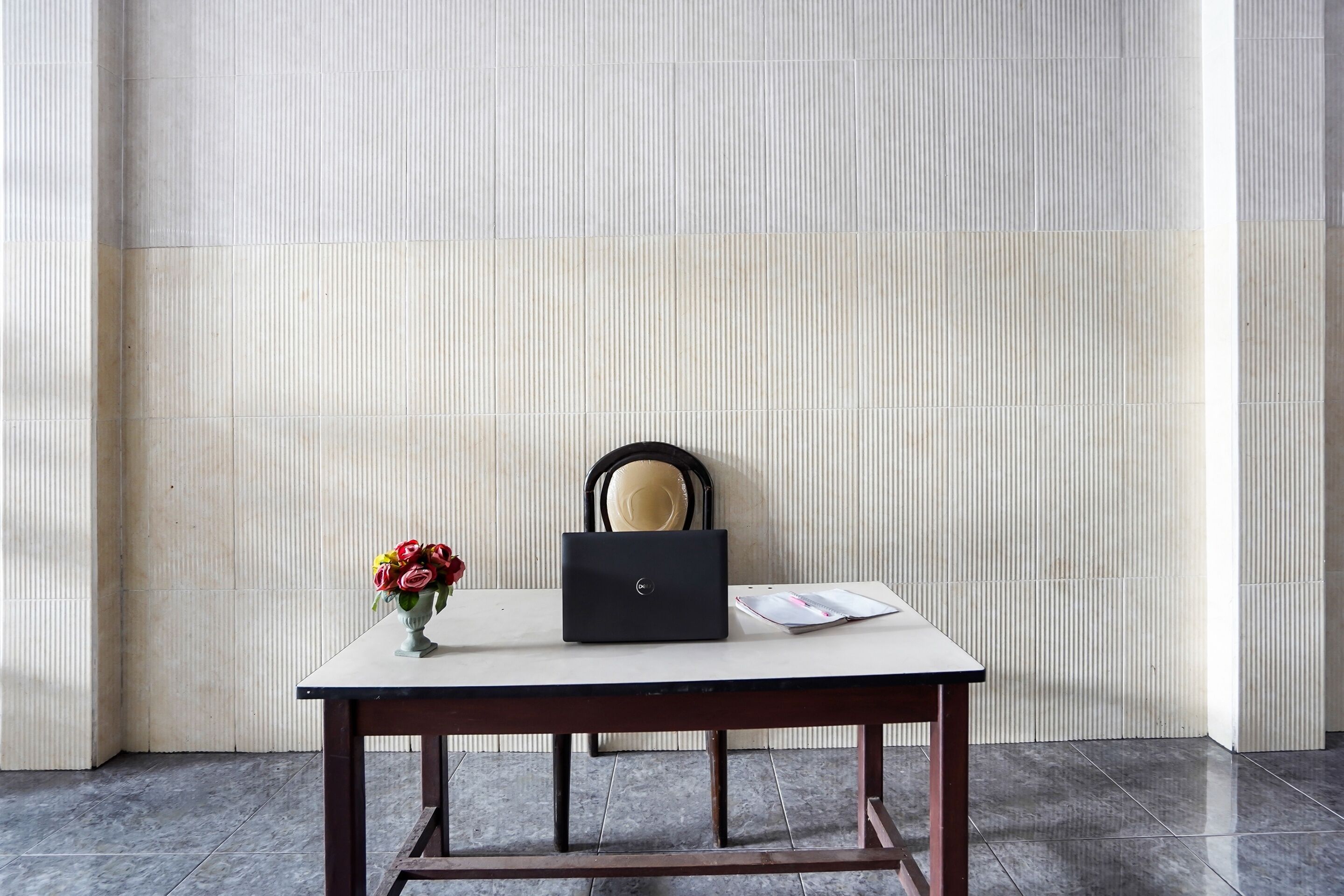 Ein Schreibtisch mit einem Laptop, einer Blumenvase und einem Stuhl vor einer gefliesten Wand.