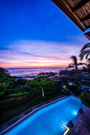 Relax by the serene pool surrounded by lush greenery and palm trees, offering a tranquil evening atmosphere.