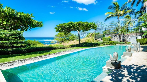 A swimming pool with clear blue water, surrounded by greenery and palm trees, with a view of the ocean. - Newly Renovated, Beachfront, 3 bed Condo on Exclusive Langosta Beach! (Tamarindo)