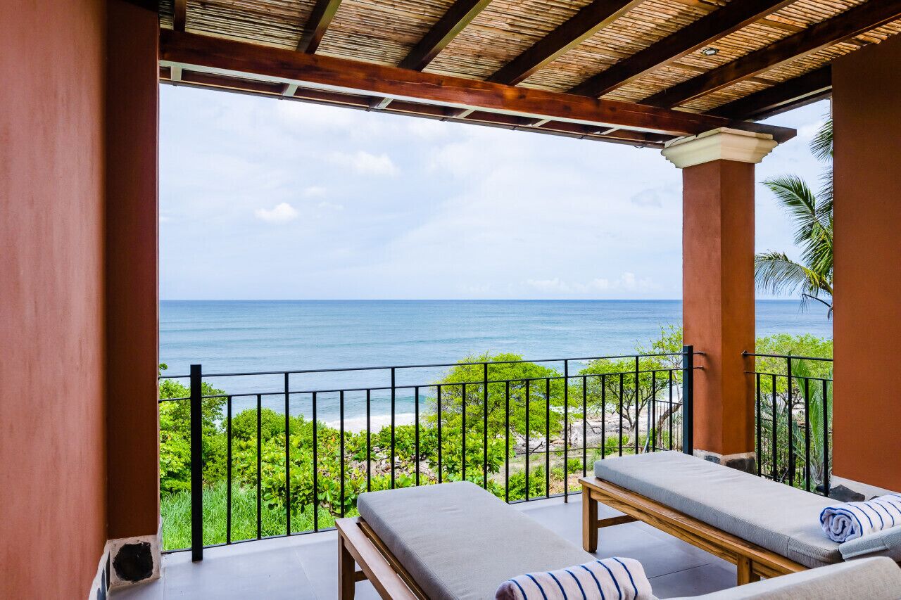 A balcony with a view of the ocean, a wooden bench, and a lounge chair with a towel.