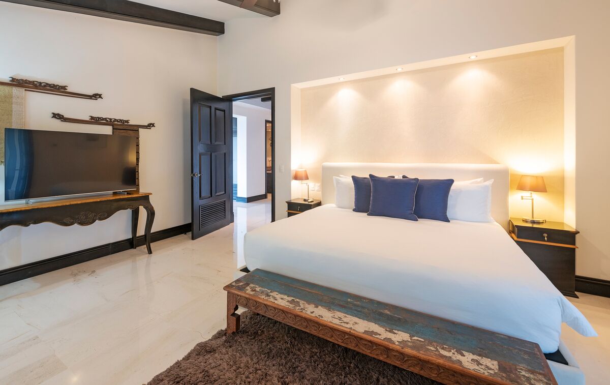 A bedroom with a large bed, bedside lamps, a TV on a wooden stand, and a door leading to another room.