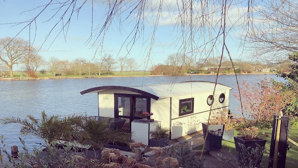 Un’elegante houseboat moderna con tetto curvo, posizionata su un lago calmo, circondata da alberi e arbusti.