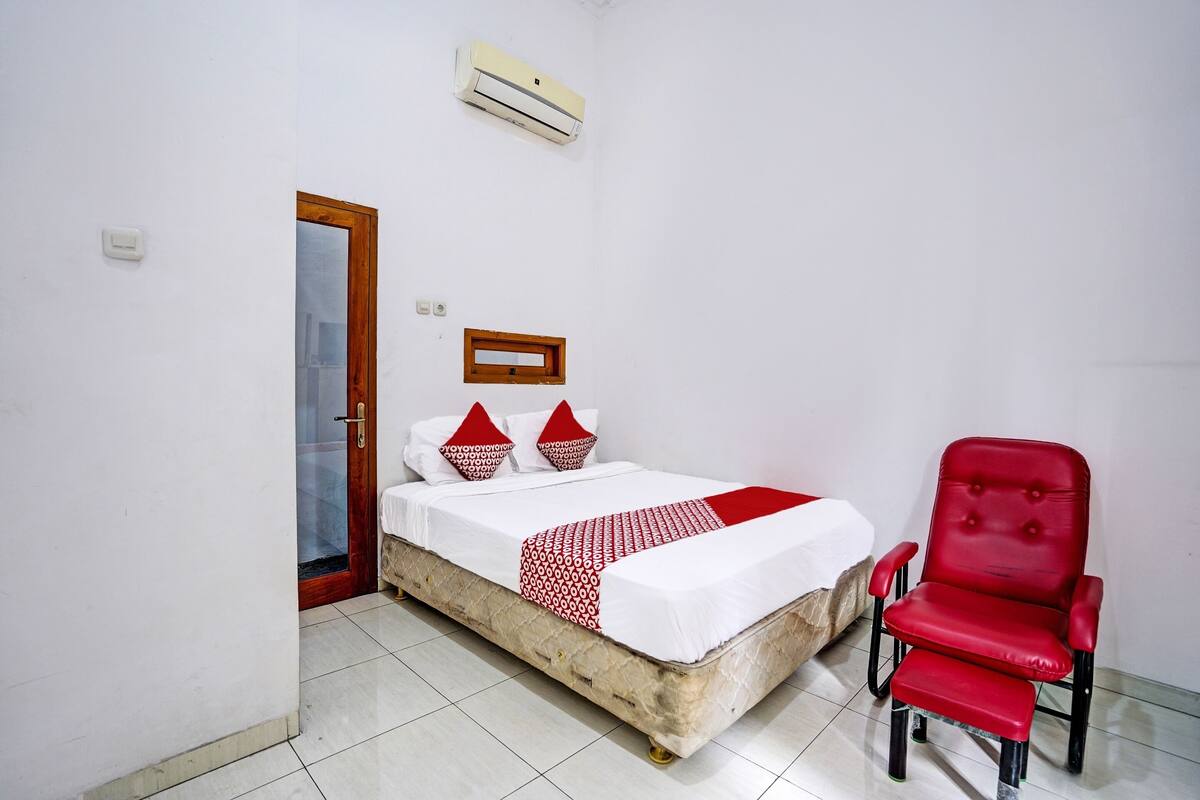 Una habitación con una cama, una silla roja, una puerta de madera y un equipo de aire acondicionado.