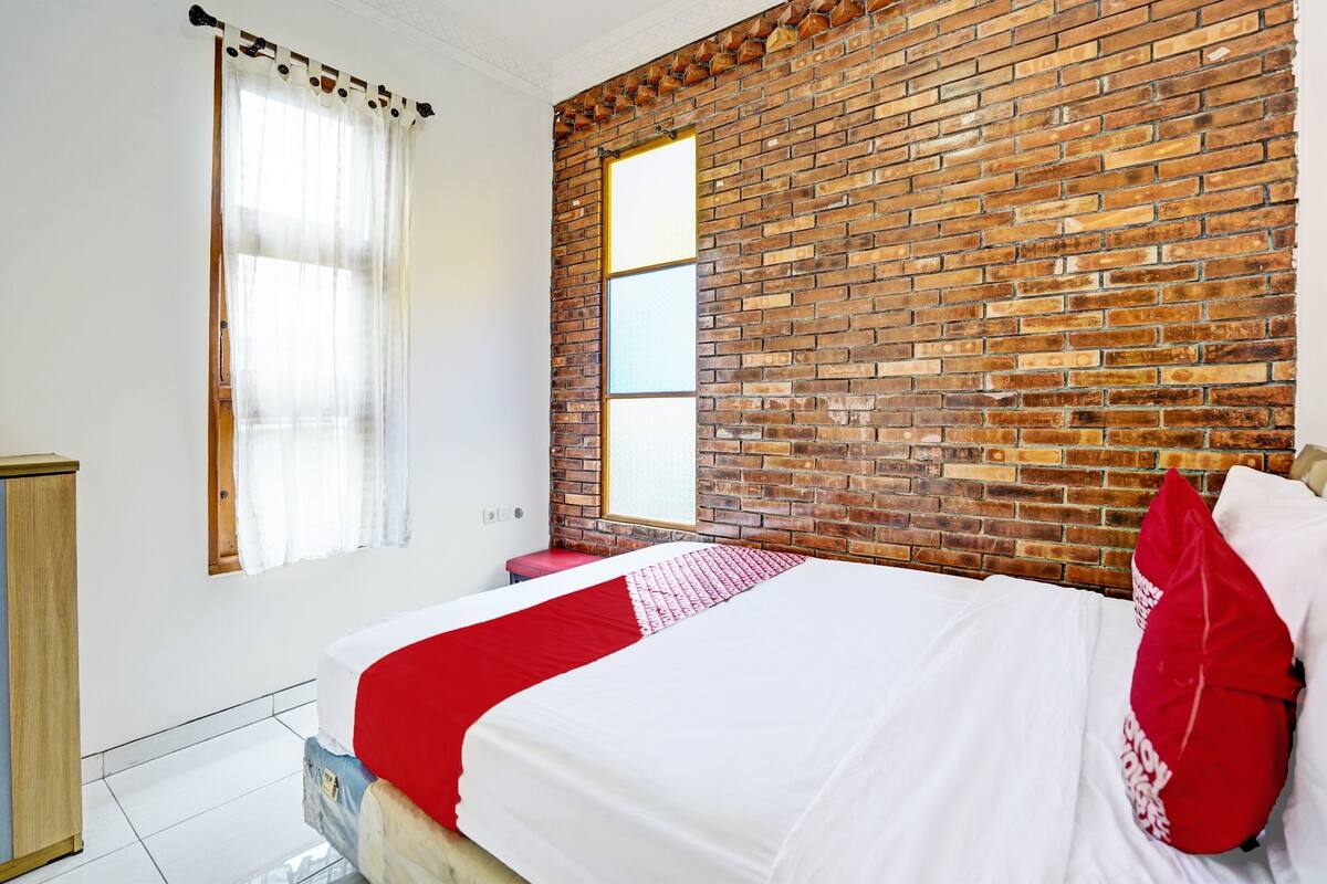 Un dormitorio con pared de ladrillo, cama con ropa de cama blanca y roja, armario de madera y ventana con cortinas traslúcidas.