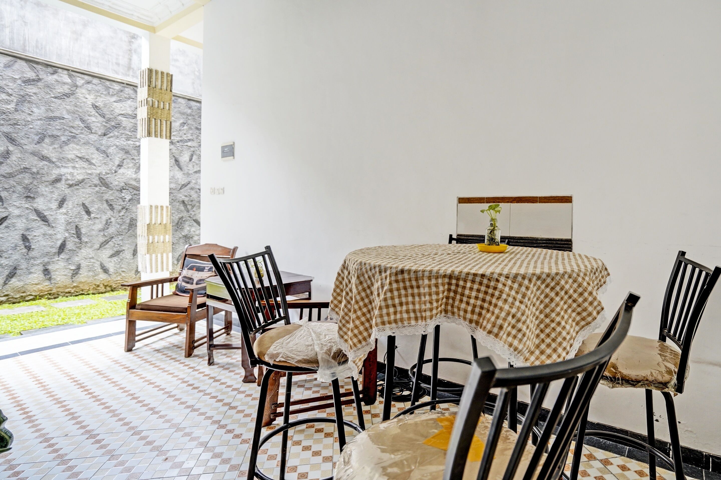 A dining area with a checkered tablecloth, wooden chairs, and a view of a garden through large windows.
