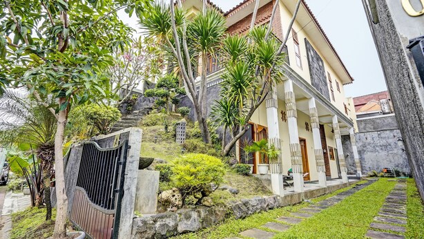 A two-story house with a balcony, surrounded by lush greenery and a stone pathway.