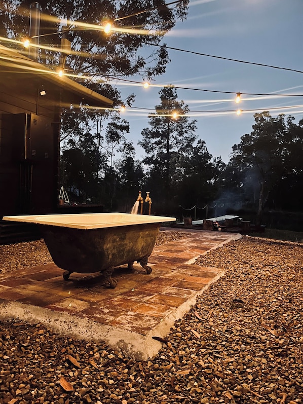 Un cadre extérieur rustique avec une grande baignoire, un sol en gravier et des guirlandes lumineuses suspendues au-dessus.