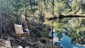 A serene outdoor setting with a swing chair by a calm body of water, surrounded by trees and natural landscape. - Little Olive Eco Cabin on a private dam.. Perfect for a relaxing weekend away! (Parklands)
