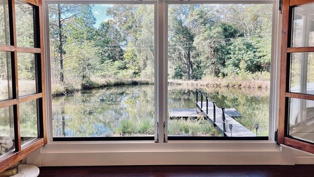A room with a large window overlooking a natural landscape, a wooden bench, and two stools. - Little Olive Eco Cabin on a private dam.. Perfect for a relaxing weekend away! (Parklands)