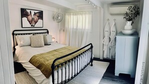 A bedroom with a bed, a bedside table, a fan, a window with curtains, and a white cabinet with a vase. - Little Olive Eco Cabin on a private dam.. Perfect for a relaxing weekend away! (Parklands)