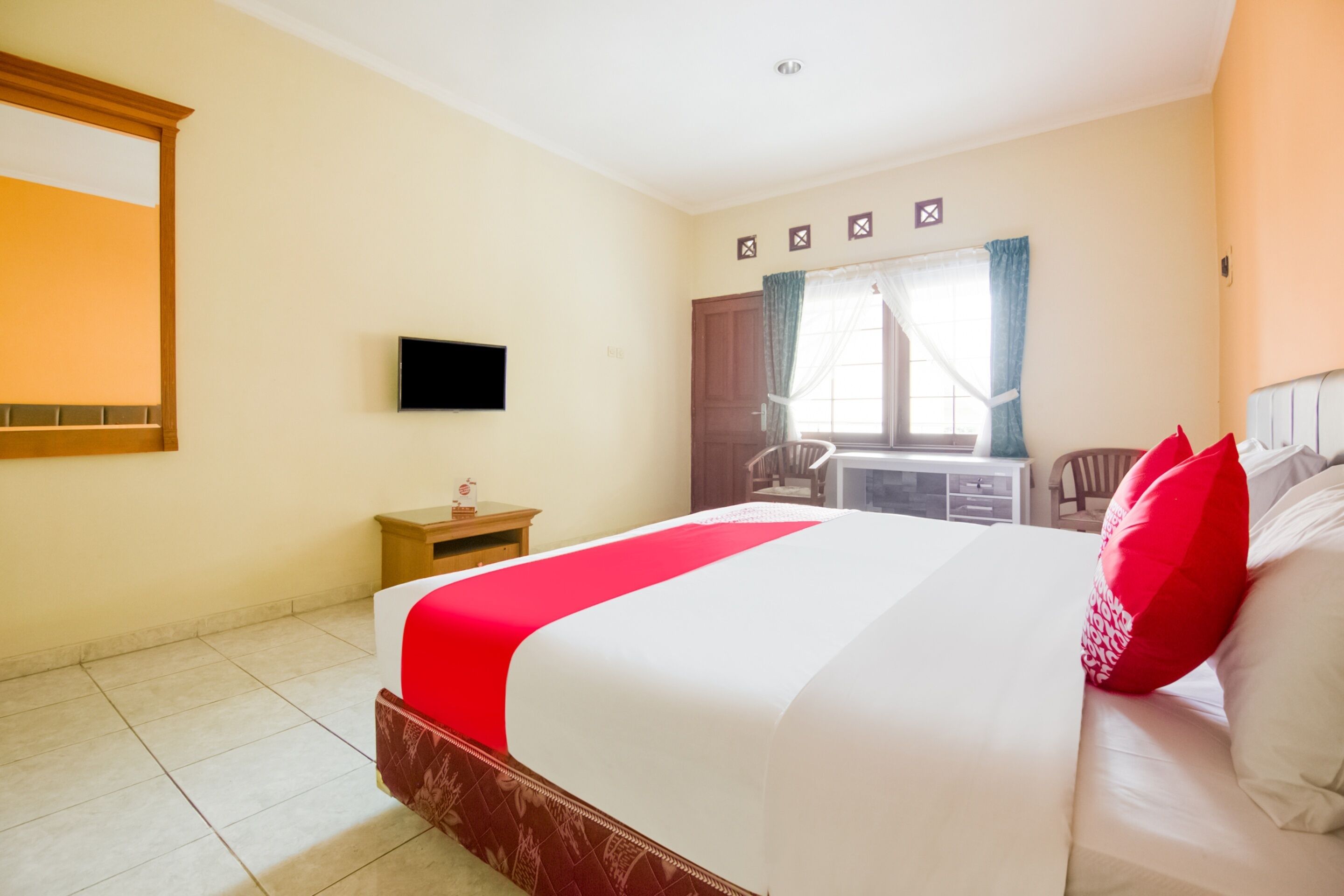 Ein Hotelzimmer mit einem großen Bett, einem an der Wand befestigten Fernseher, einem kleinen Tisch mit einem Stuhl und einem Fenster mit Vorhängen.