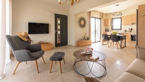 Een moderne woonkamer met een grijze bank, een bijpassend voetenbankje en een salontafel. Er is een televisie aan de muur bevestigd en er is een keukenruimte zichtbaar door een open deur.
