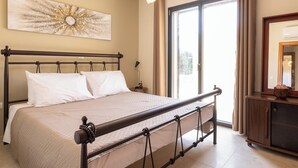 Een net opgemaakt bed met hoofdbord, een nachtkastje met lamp, een groot raam met gordijnen en een aan de muur hangend kunstwerk.