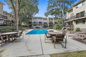 A pool area with lounge chairs and tables, surrounded by trees and a multi-story building. - 302 Tanglewood Villa (Hilton Head Island)