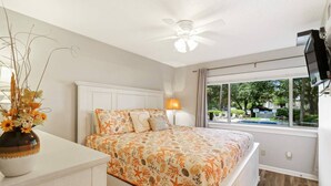 Un dormitorio con cama, mesita de noche, florero con flores, ventilador de techo y ventana con vistas al exterior.