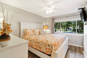 A bedroom with a bed, a nightstand, a vase with flowers, a ceiling fan, and a window with a view of the outdoors. - 302 Tanglewood Villa (Hilton Head Island)