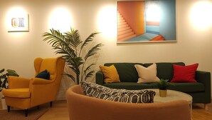 A living room with a yellow armchair, a green sofa with multiple cushions, a large potted plant, and a painting of a staircase. - Acro Capsule Hotel Namba Dotonbori (Osaka)