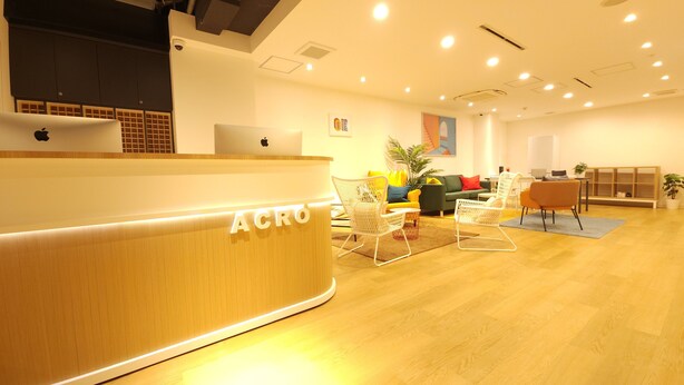 A modern reception area with a wooden desk labeled "ACRO," a seating area with colorful chairs, and a plant. - Acro Capsule Hotel Namba Dotonbori (Osaka)