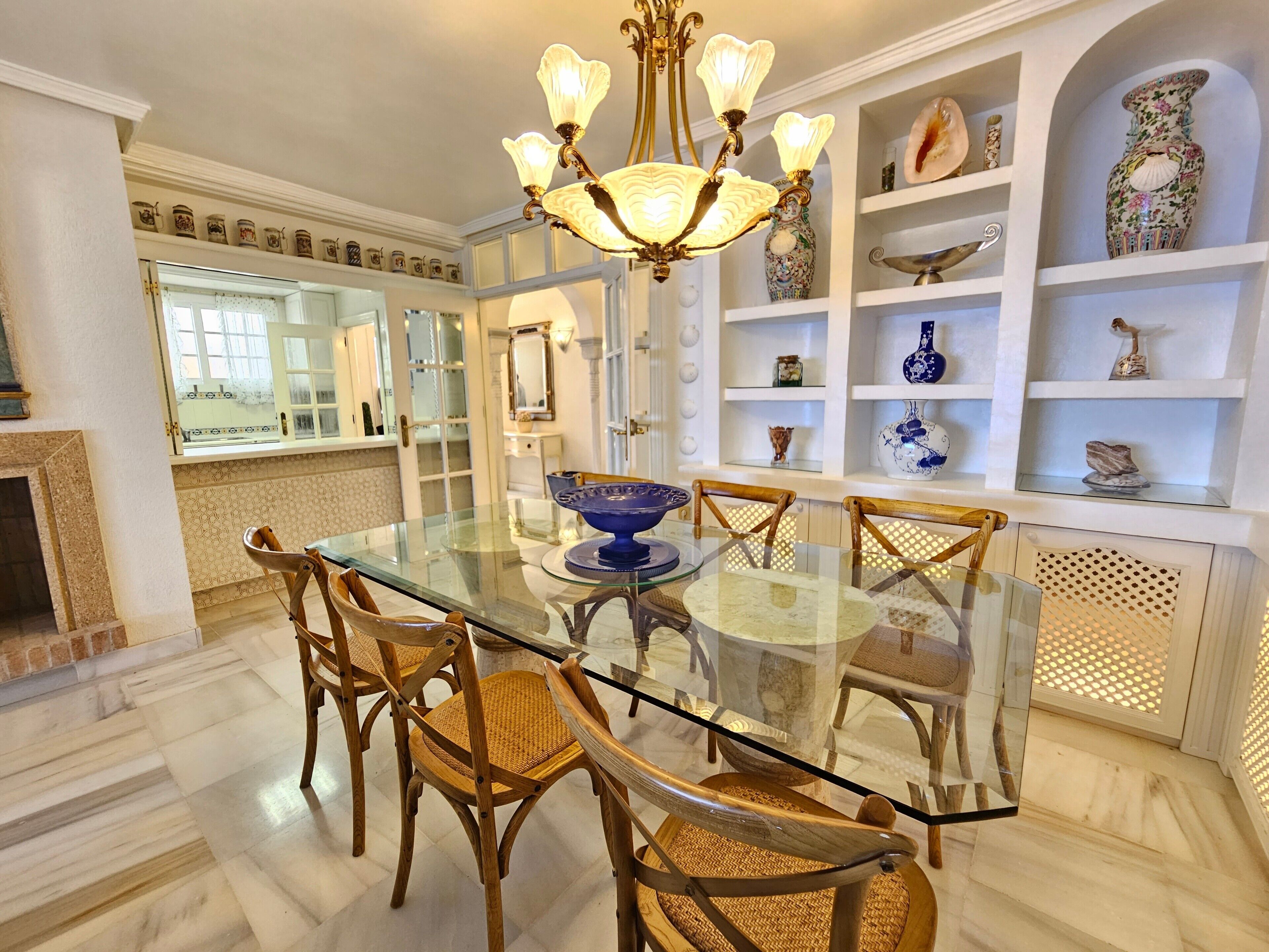 Een eethoek met een glazen tafel, rieten stoelen, een kroonluchter en decoratieve schappen.