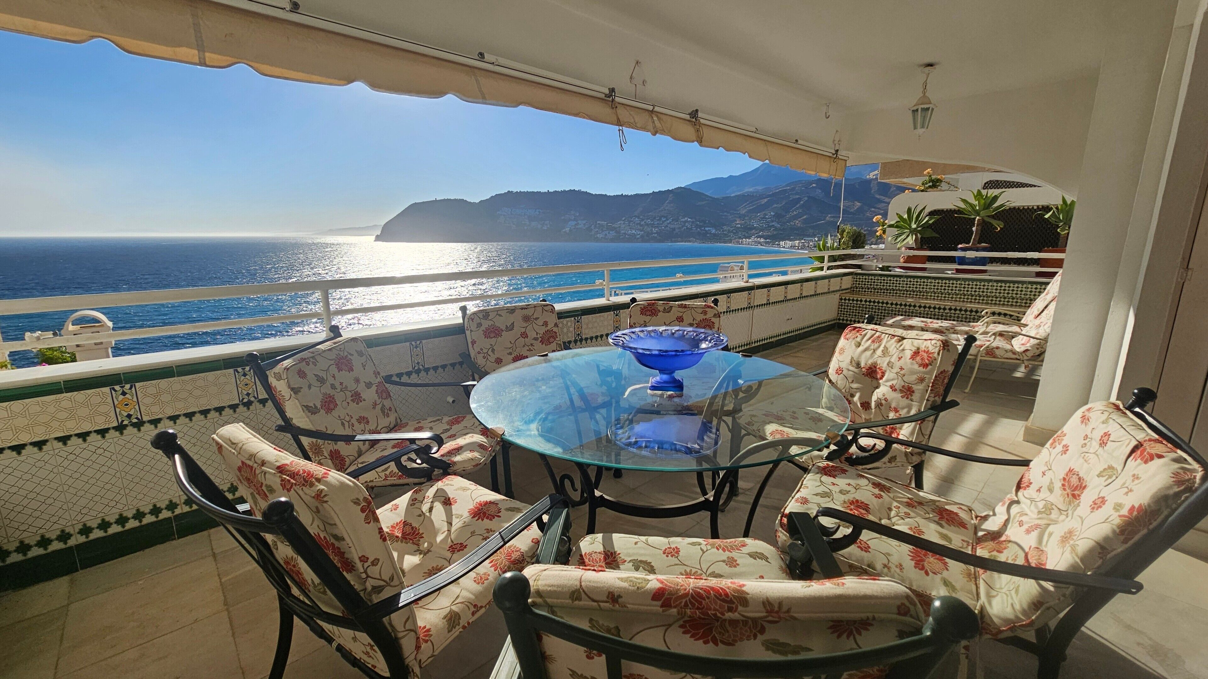 Een balkon met een glazen tafel, bloemenstoelen en uitzicht op zee en bergen.