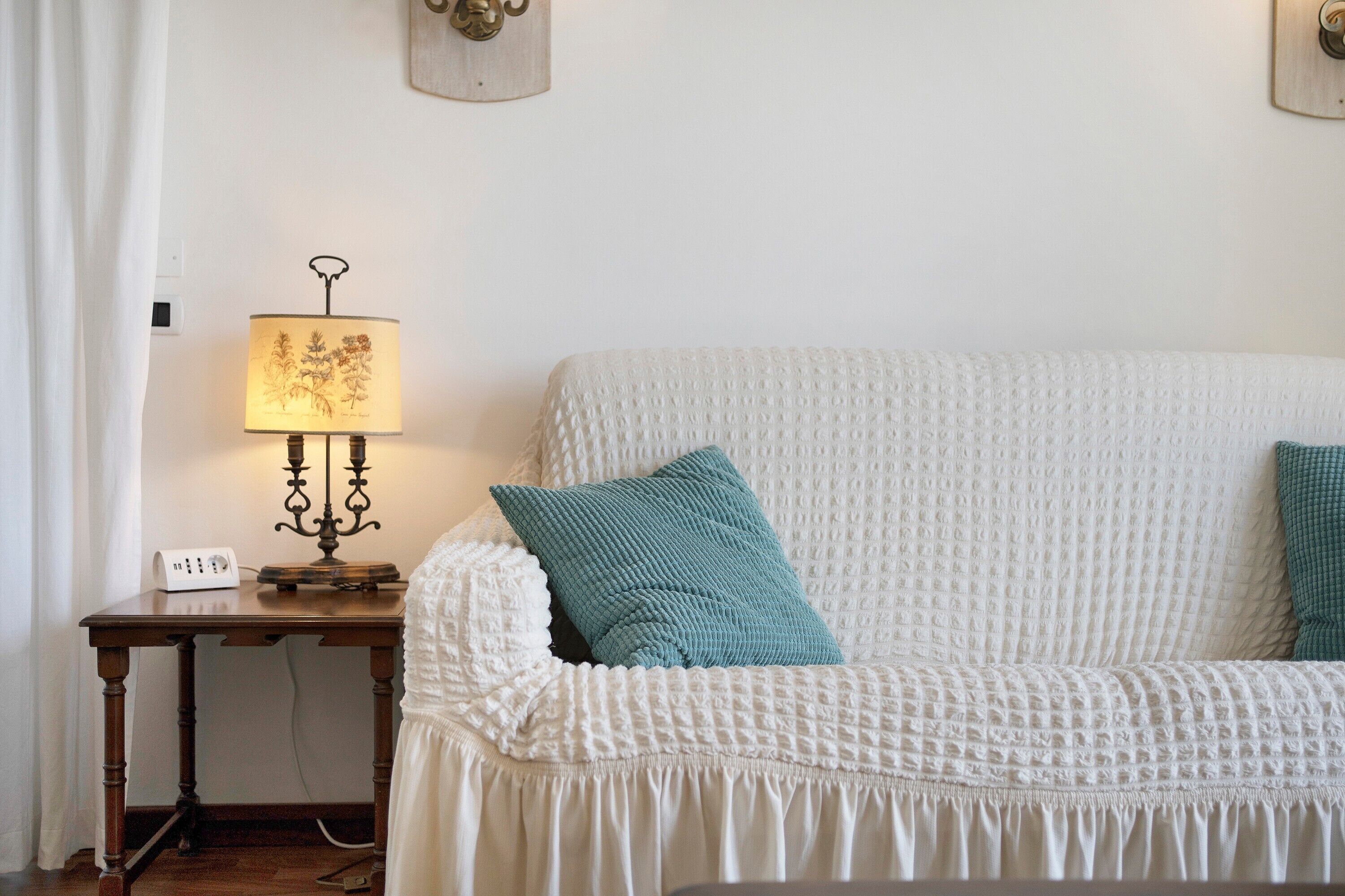 Een witte gestructureerde bank met blauwgroene kussens, een houten bijzettafeltje met een lamp en een stekkerdoos.