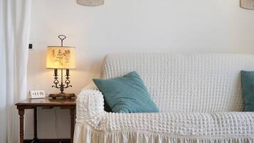 Een witte gestructureerde bank met blauwgroene kussens, een houten bijzettafeltje met een lamp en een stekkerdoos.