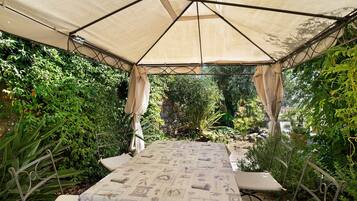 Eine Außenpatio mit einem Tisch, der mit einer gemusterten Tischdecke bedeckt ist, umgeben von Metallstühlen, unter einem Baldachin.