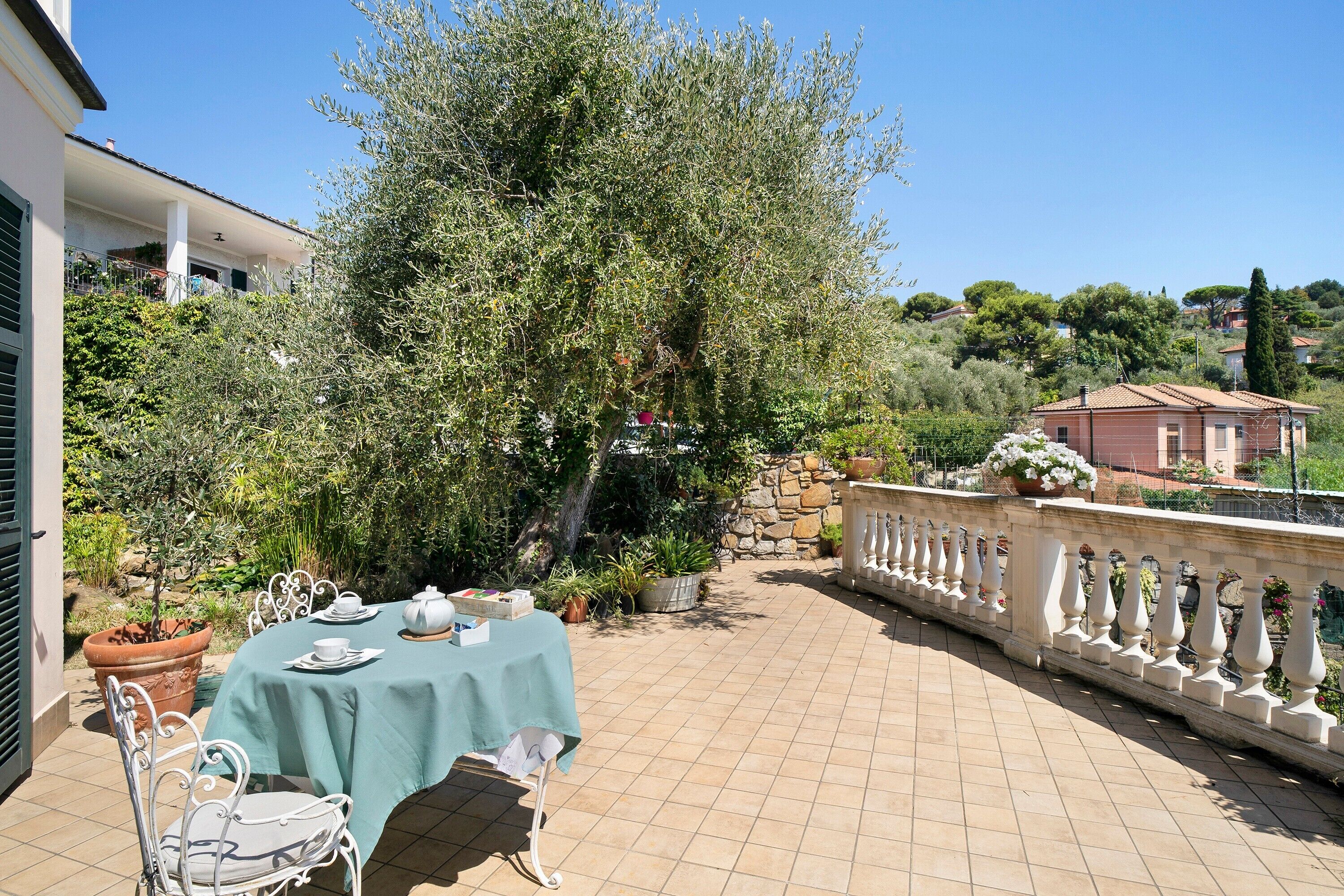 Eine Terrasse mit Tisch für zwei Personen, umgeben von Topfpflanzen und einem großen Baum.