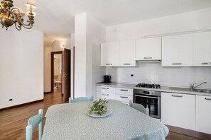 Une cuisine moderne avec des meubles blancs, une cuisinière et un évier. On y trouve une table à manger recouverte d’une nappe à motifs floraux, ainsi que des chaises.