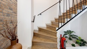 Un escalier avec des marches en bois et une rampe métallique, un mur de briques, un tableau encadré, un vase avec des branches et un extincteur.