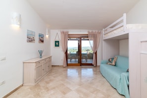 Une pièce comprenant un lit superposé, un canapé, une armoire, une porte-fenêtre coulissante donnant sur une terrasse et des œuvres d’art accrochées au mur.