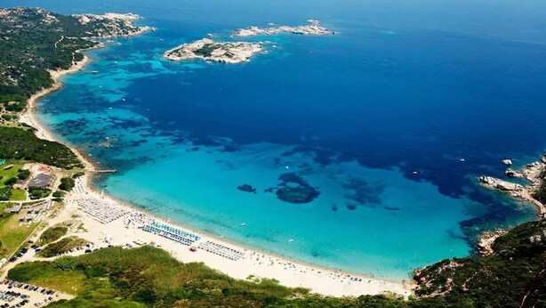An aerial view of a coastal area with a beach, clear blue waters, and rocky outcrops.