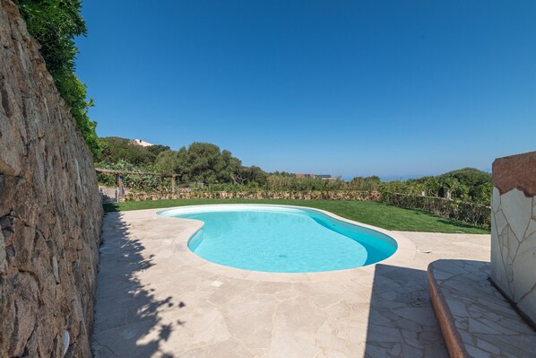 Una piscina de borde curvo, rodeada de baldosas de piedra y un muro de piedra.