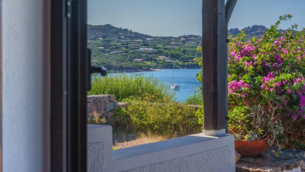 A coastal view from a covered outdoor area with a stone wall, potted plants, and a clear sky.