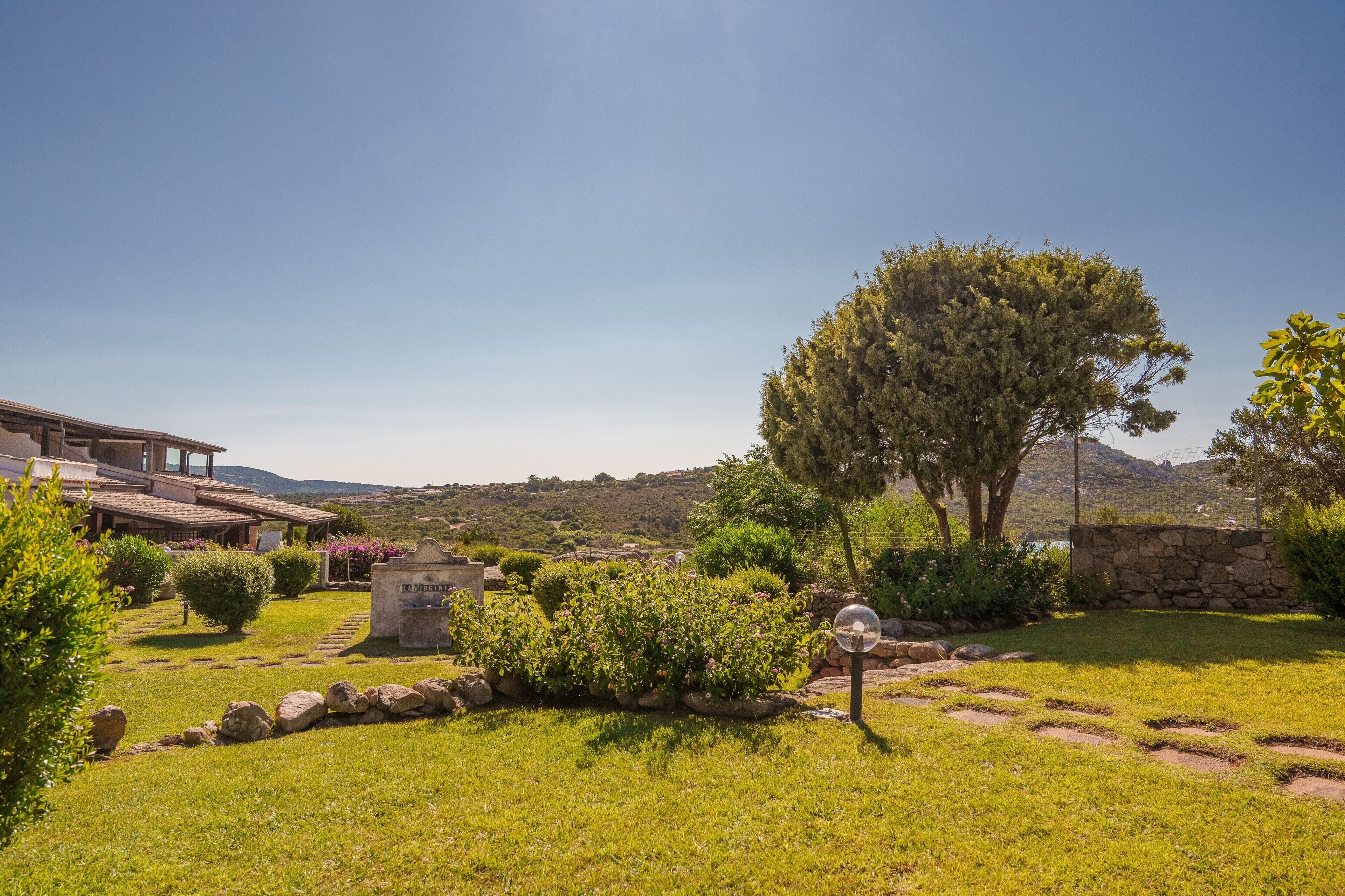 Un jardín ajardinado con un muro de piedra, un árbol grande y un edificio con techo inclinado.