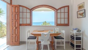 A dining area with a wooden table, white chairs, a view through a window, and a white shelf with baskets. - Apartment "Kind of Blue" with Sea View, Wi-Fi and Air Conditioning (Siracusa)