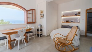 A bedroom with a bed, a desk with chairs, a window with a view, and a shelf with items. - Apartment "Kind of Blue" with Sea View, Wi-Fi and Air Conditioning (Siracusa)