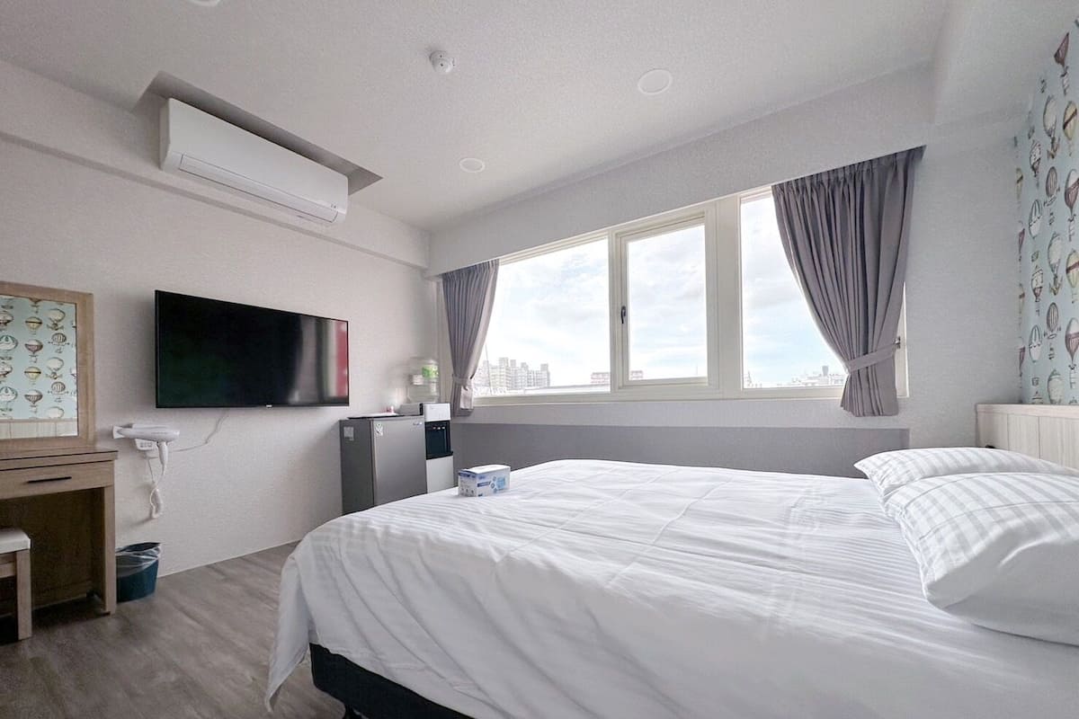 Quarto de hotel com cama, escrivaninha, televisão e janela com cortinas.