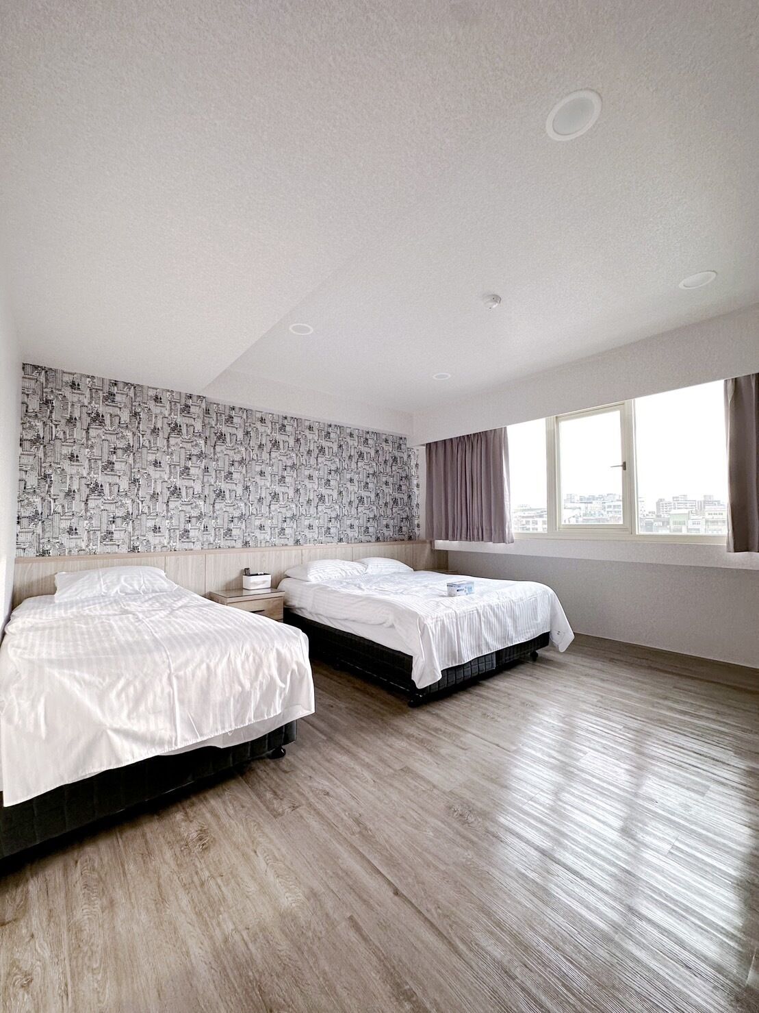 A hotel room with two beds, a patterned wallpaper, a large window with curtains, and a wooden floor.