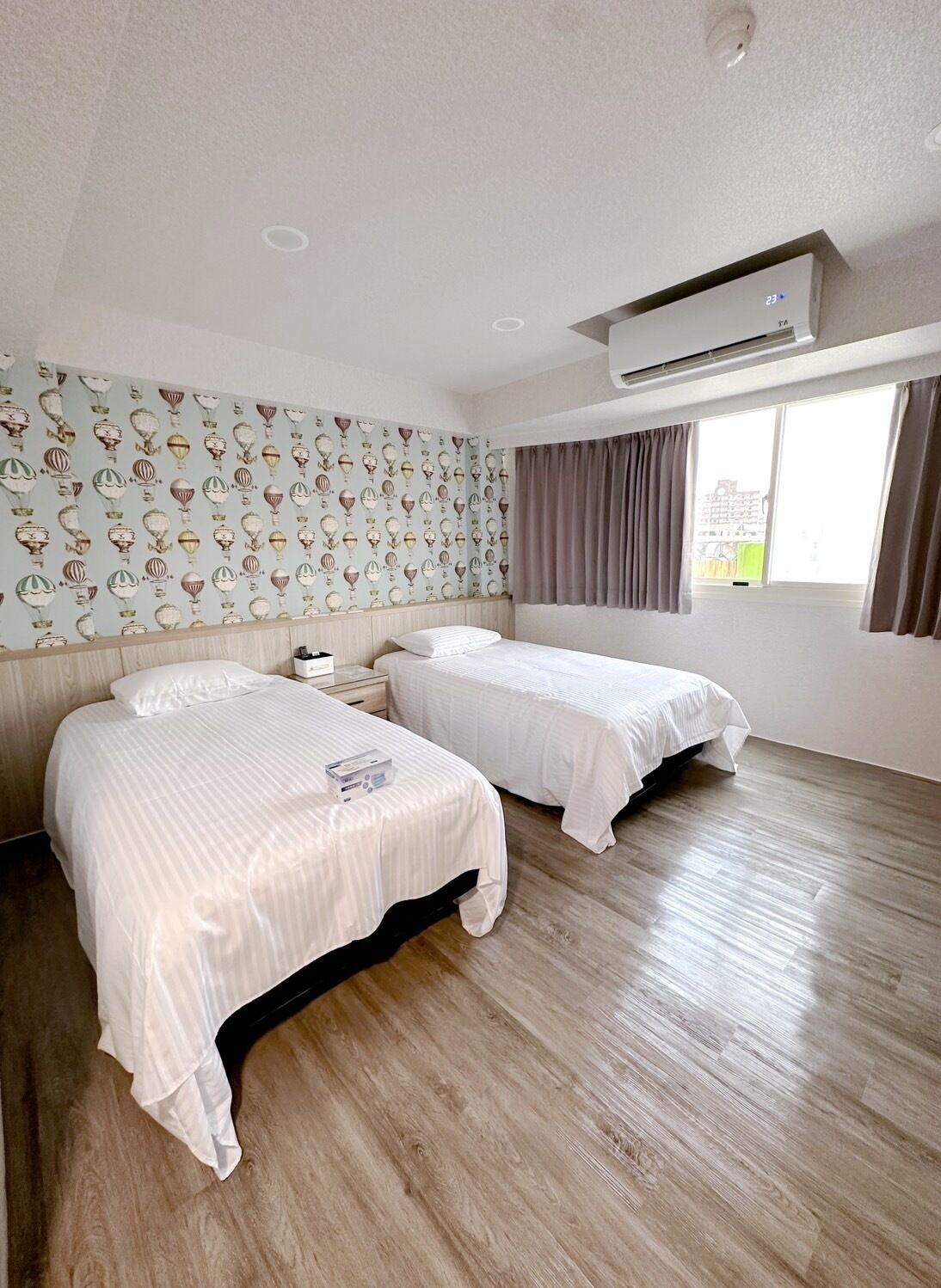 A hotel room with two beds, a patterned wallpaper, a window with curtains, and an air conditioning unit.