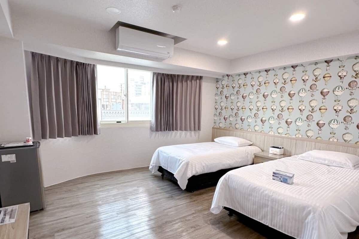 Quarto de hotel com duas camas, uma escrivaninha, um armário cinzento e uma janela com cortinas.