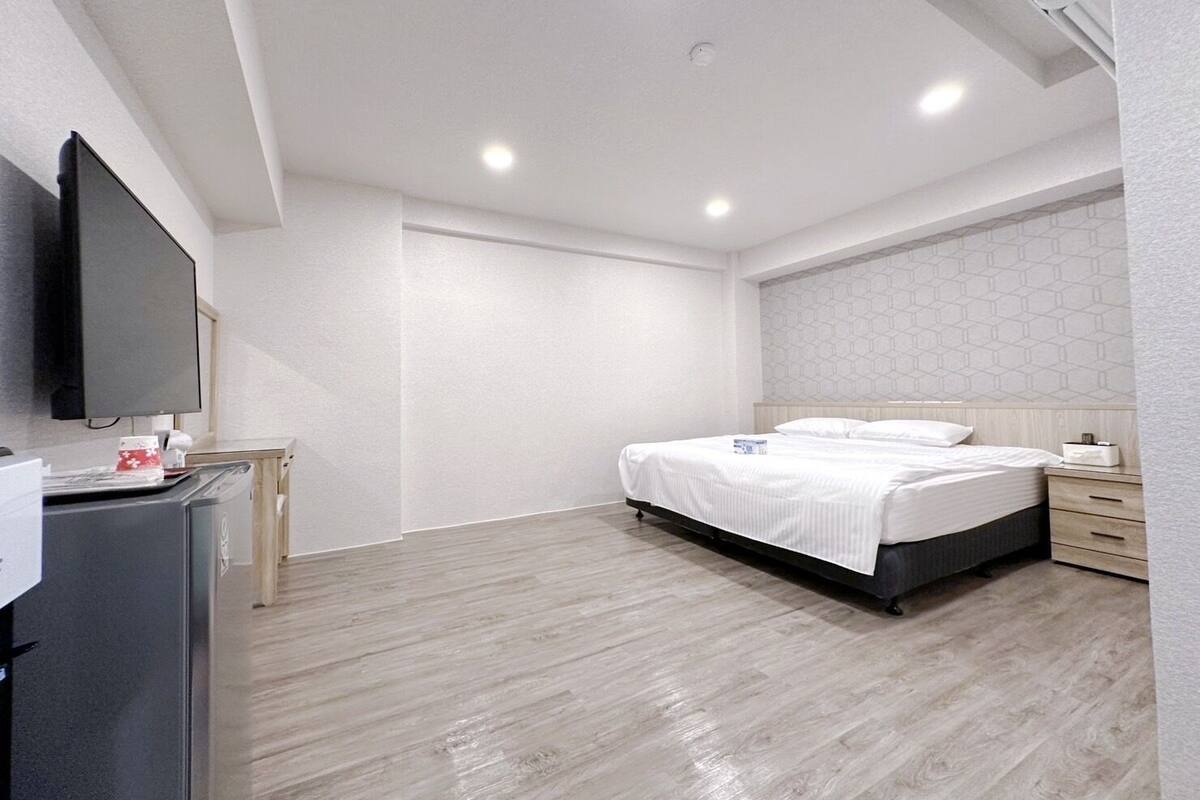 Quarto de hotel moderno com uma cama grande, uma TV de tela plana fixada na parede, piso de madeira e parede branca com padrão geométrico.