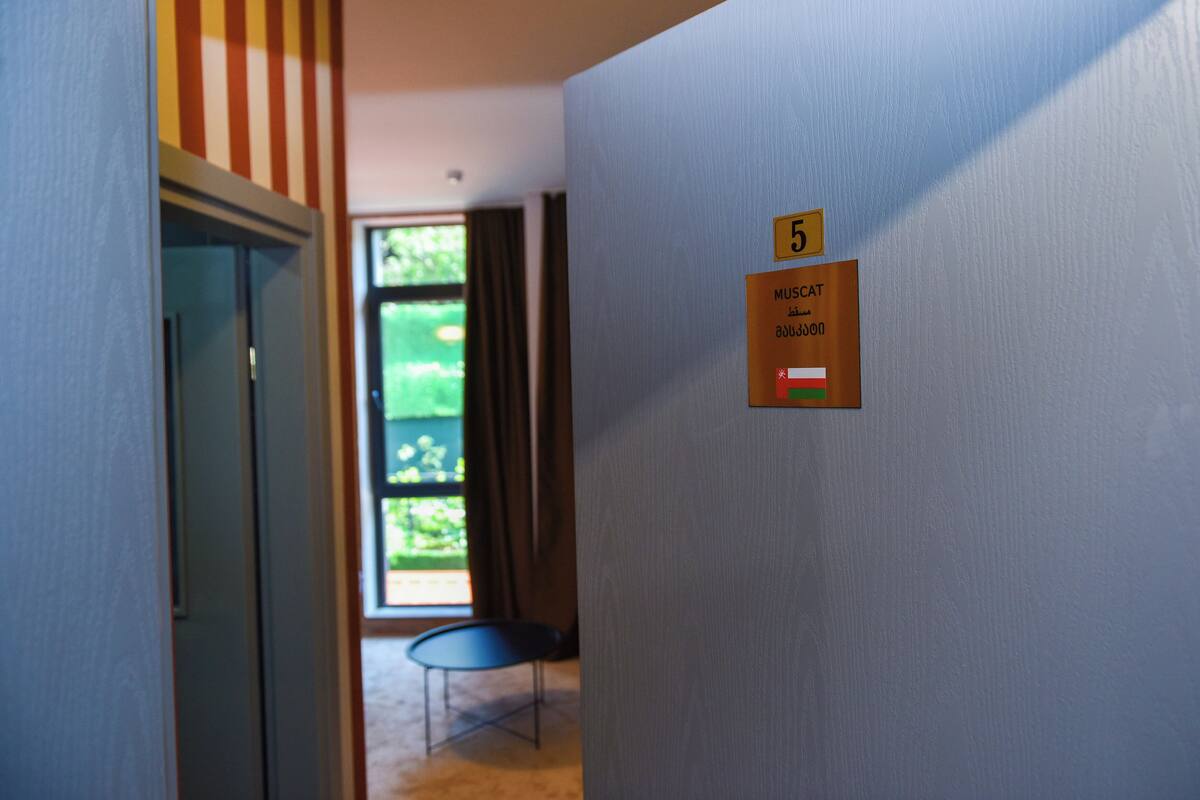 Une chambre d’hôtel avec une porte, une fenêtre avec des rideaux, une petite table et une plaque sur le mur indiquant le numéro de chambre : 5.
