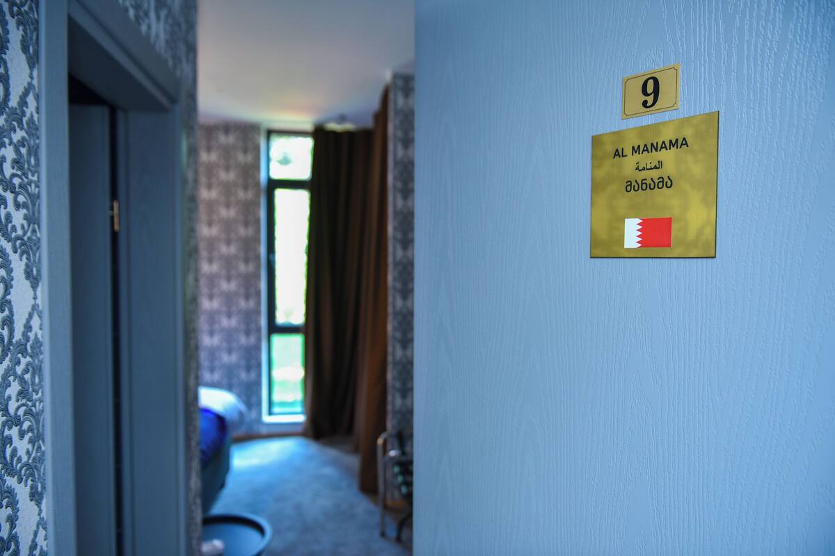 Un couloir avec une porte à gauche et un mur sur lequel est affiché un panneau indiquant le numéro de la chambre 9, Al Manama, ainsi qu’un drapeau.