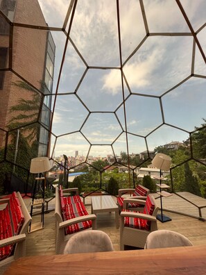 Een terras op het dak met honingraatvormige glazen panelen, rieten meubels en uitzicht over de stad.