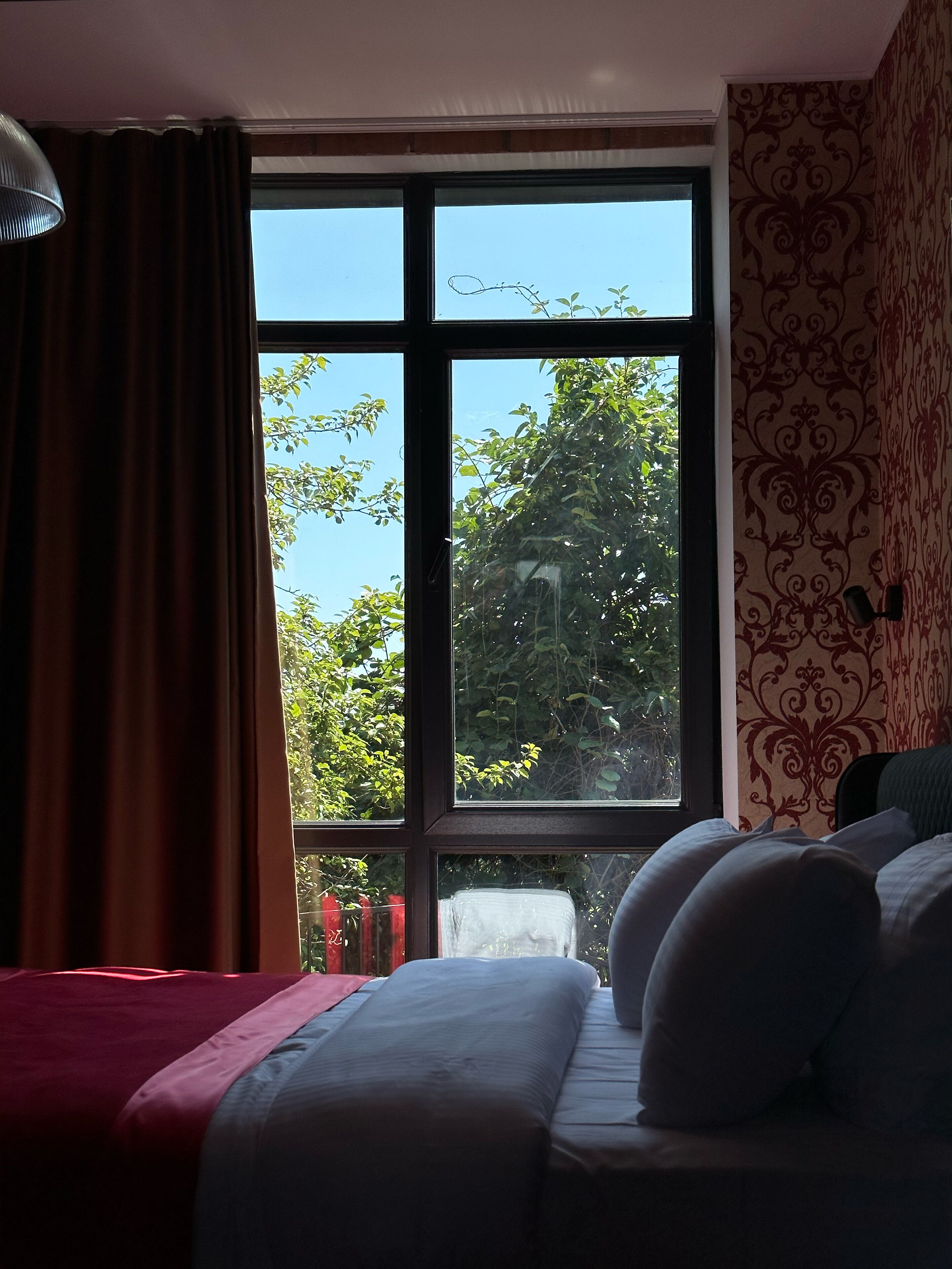A bedroom with a large window, a bed with pillows, a lamp, and curtains.