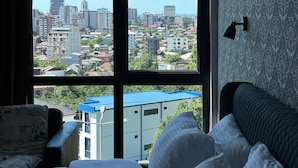 Une chambre d’hôtel dotée d’une grande fenêtre donnant sur la ville, d’un lit avec une literie blanche, d’une tête de lit sombre et d’une lampe fixée au mur.