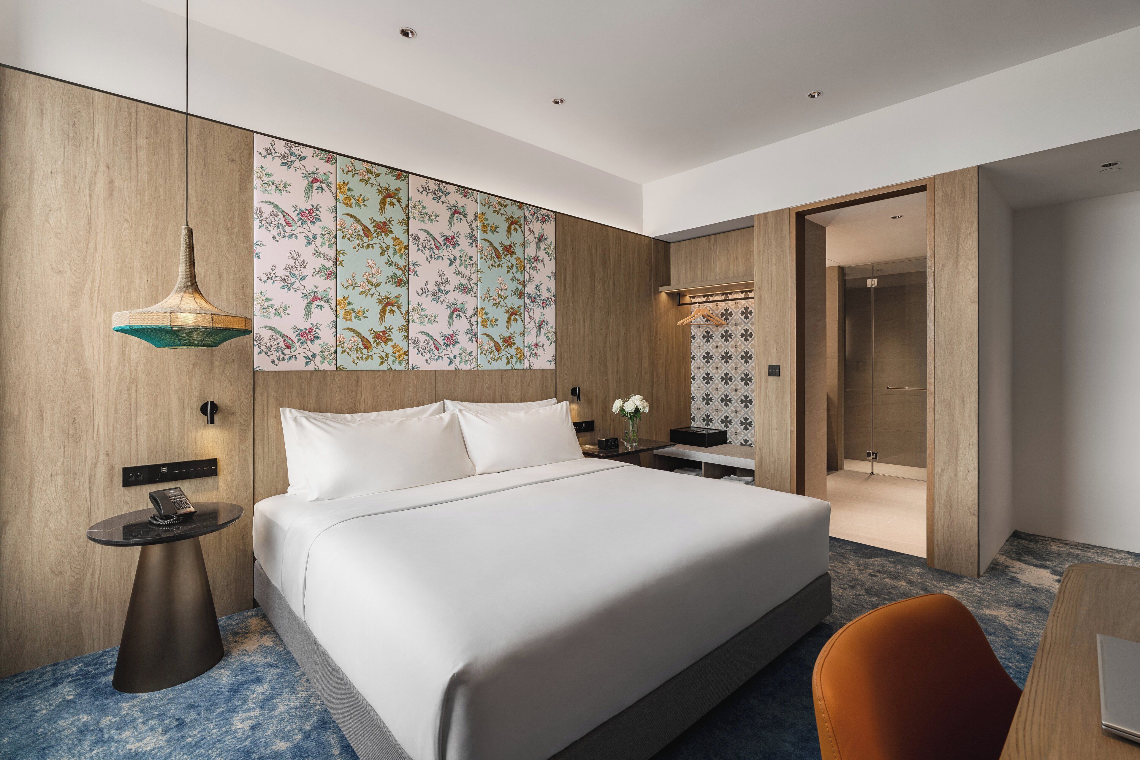 A modern hotel room with a large bed, a round side table with a phone, a hanging light fixture, and a decorative wall panel.